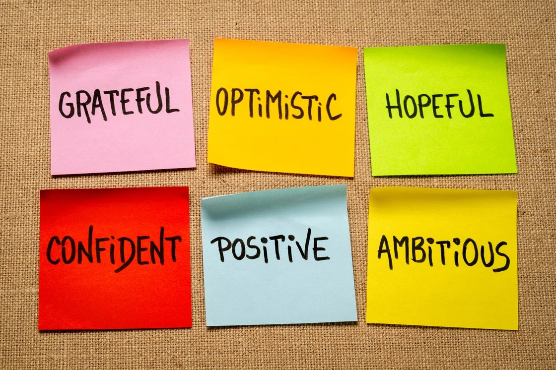 Colorful sticky notes with positive adjectives: grateful, optimistic, hopeful, confident, positive, ambitious on cork board